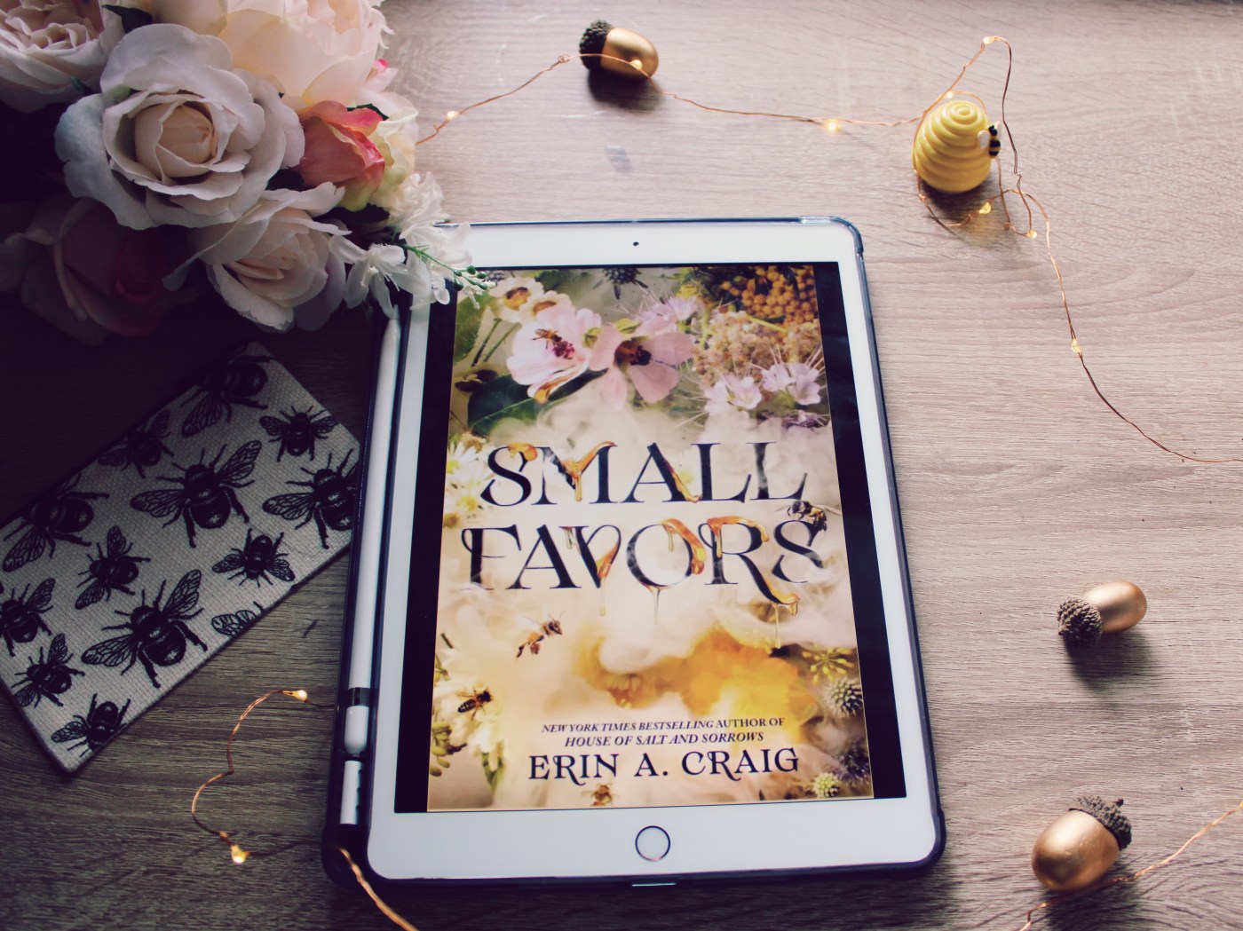 Small Favors book cover surrounded by acorns, beehive, and bee quarter bag. Pink flowers in upper left corner, as well as fairy lights decorate the rest of the scene.