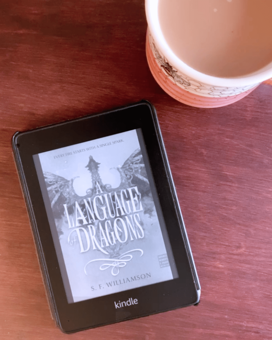 kindle sitting on a dark cherry brown coffee table showing the cover of language of dragons with a cup of tea in the upper right hand corner.