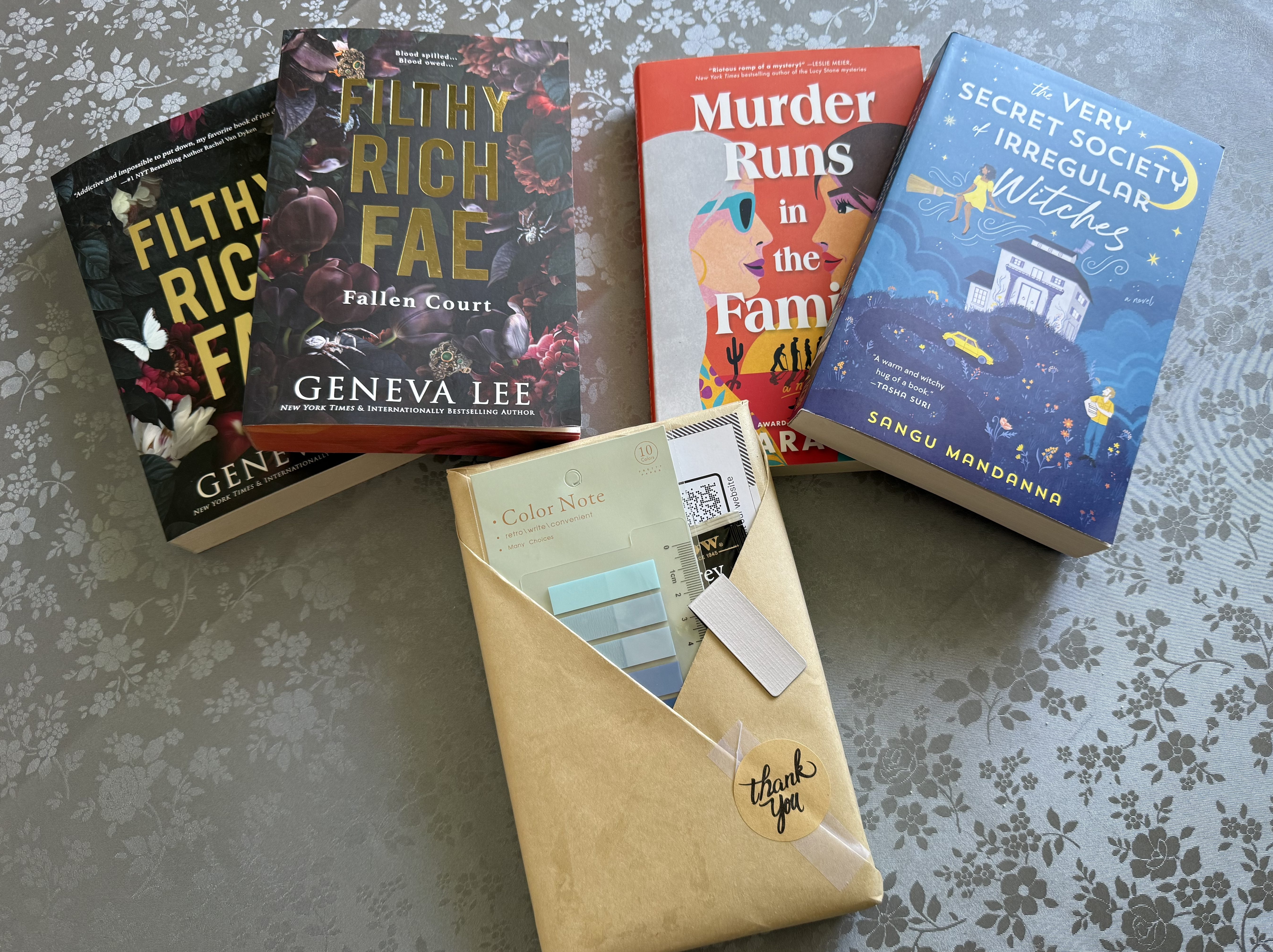 Five books laying on a blue floral table cloth, Filthy Rich Fae books on and two by Geneva Lee, Murder Runs in the Family by , and The Very Secret Society of Irregular Witches by Sangu Mandanna. The final book is still wrapped in brown paper with tabs and a bookmark tucked into the trianglar wrapping.