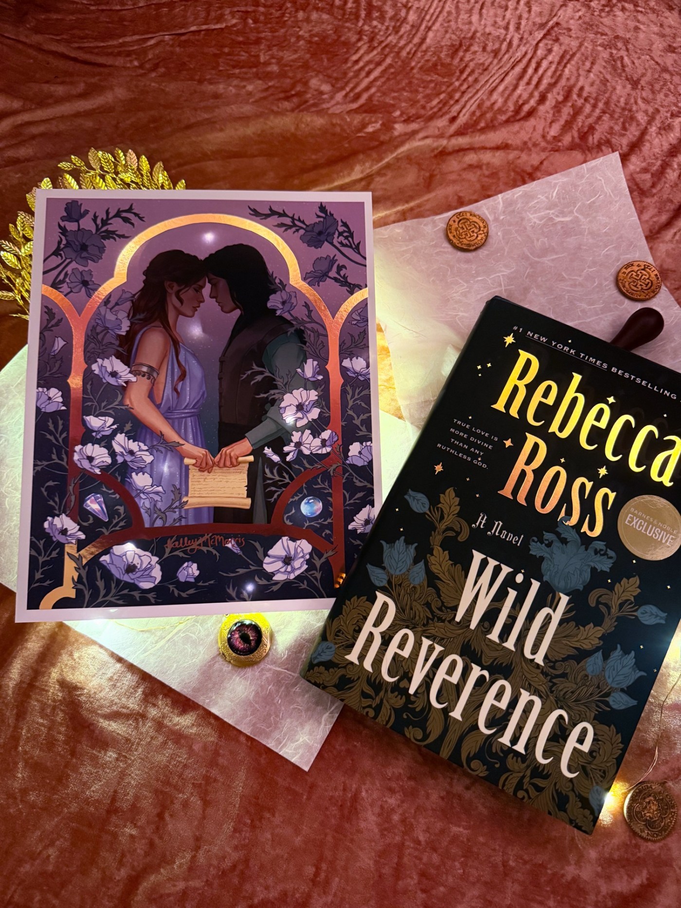 The Barnes and Noble edition of Wild Reverence as well as a print of the artwork featured on the Waterstones edition of Wild Reverence sit on a bed of dust pink velvet with doubloons and a locket with a purple sparkly eye like piece of art on it.