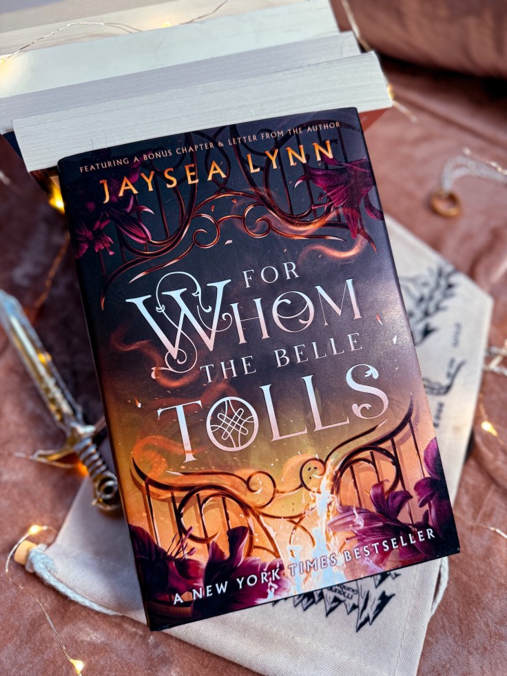 The Barnes and Noble edition of For Whom the Belle Tolls by Jaysea Lynn is resting against a pile of paperback books, pages facing out with a minature golden sword, map of middle earth, and the one ring surrounding / underneath.