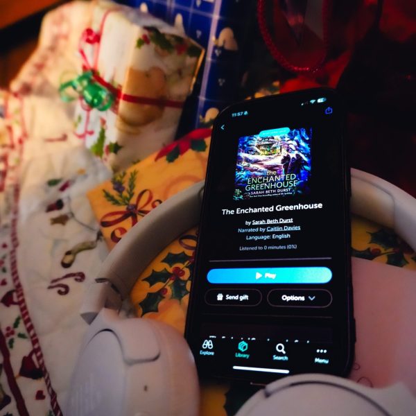 phone sitting on a pile of christmas presents and a pair of white headphones showing the libro.fm version of the enchanted greenhouse audiobook.