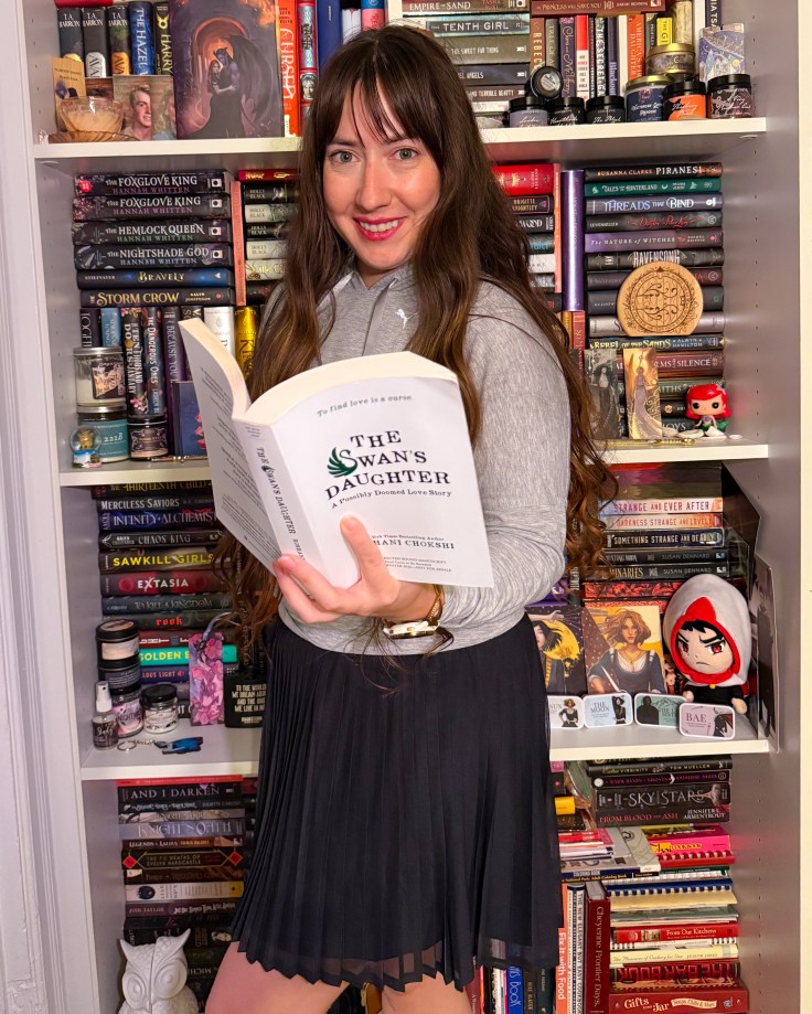 Kaite standing in front of her bookshelves holding the bound manuscript of The Swan's Daughter