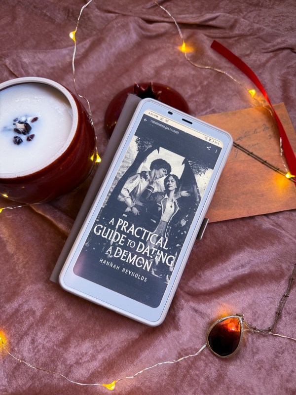 Ereader with a Practical Guide to Dating a Demon on the screen laying on a pink velvet background with a pomegranate candle and a necklace with a string of lights around it.