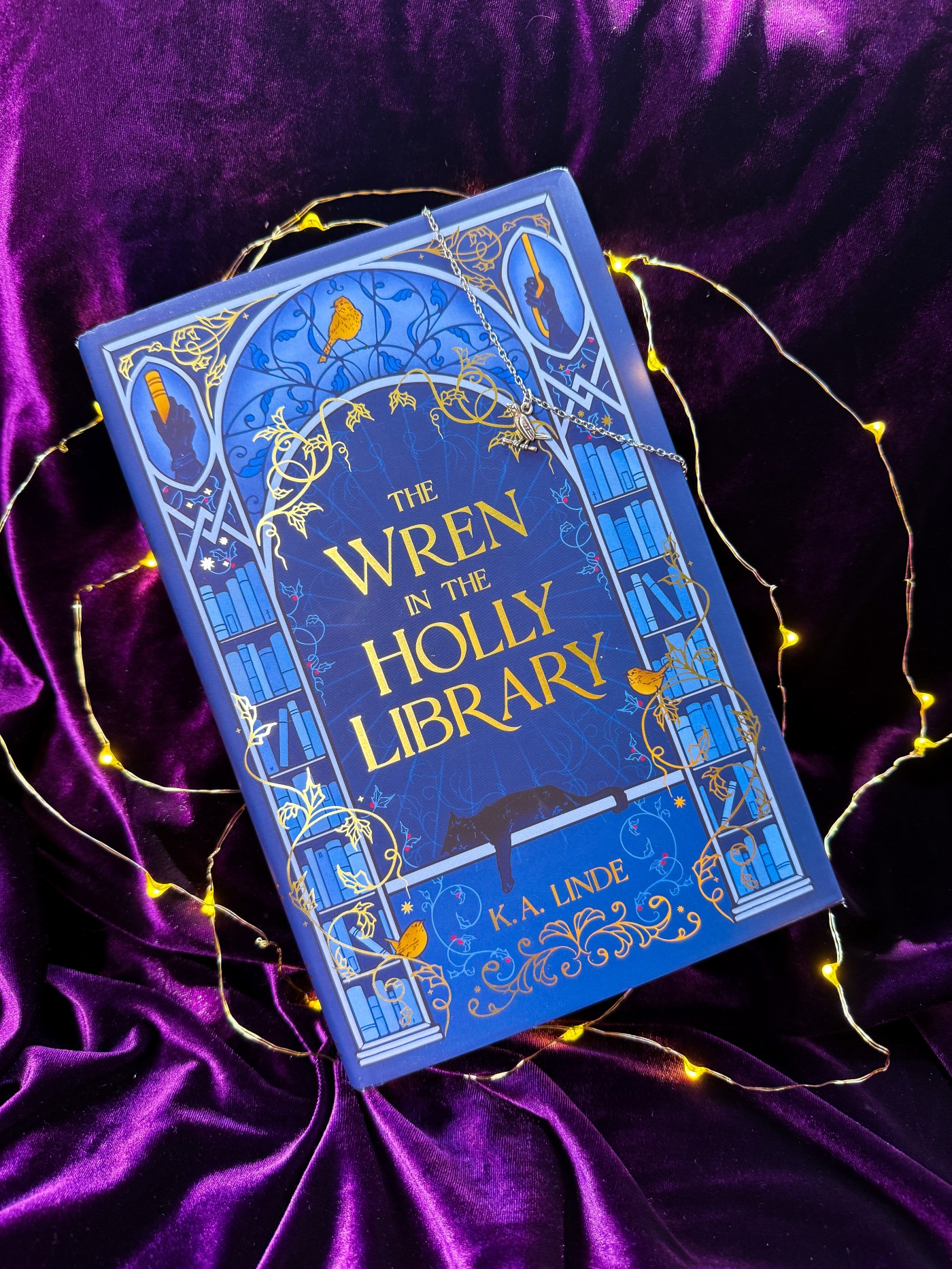 The Fairyloot edition of the Wren in the Holly Library on a bed of purple velvet with fairy lights and a wren necklace hanging down on the cover.