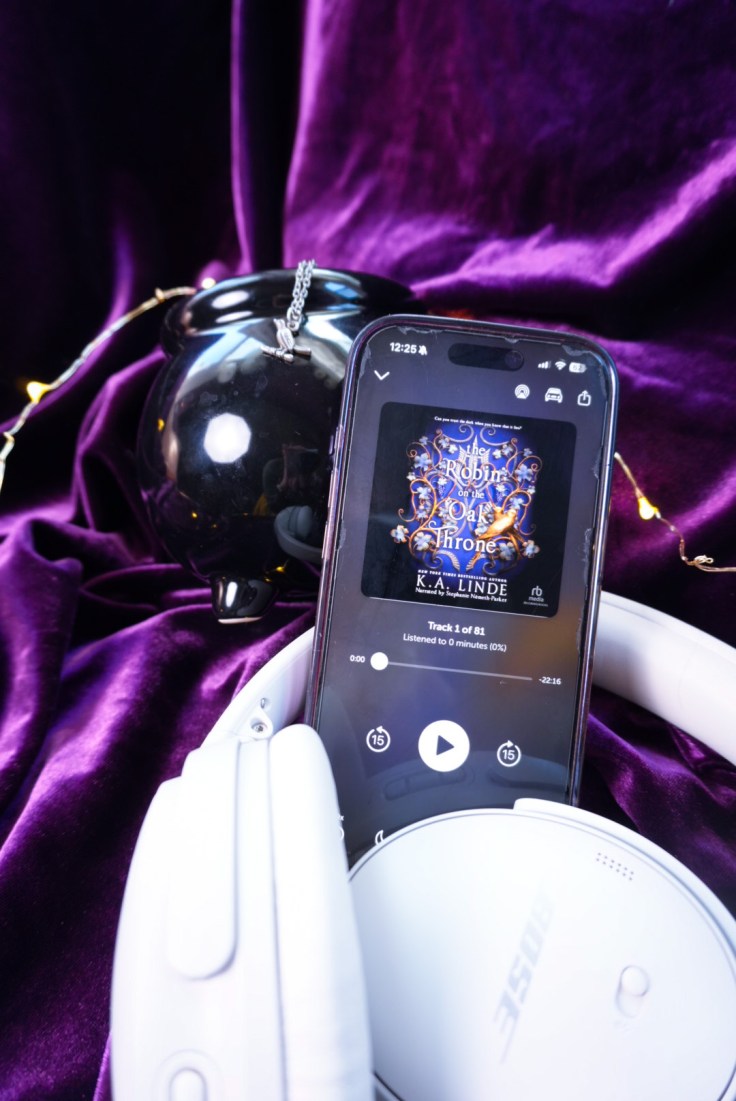 a black cauldron, with a wren necklace on it surrounded by fairly lights with an iphone showing the audiobook of the Robin on the Oak Throne and a pair on headphones, all laying on purple velvet.