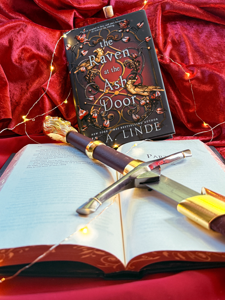 The Raven at the Ash Door book sitting on red velvet with a sword pulled out partially on top of it surrounded by twinkle lights.
