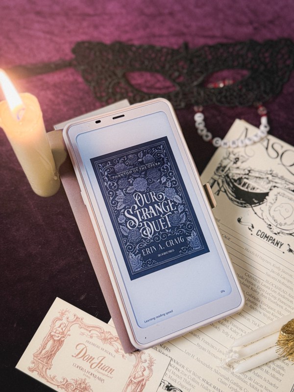 Boox Palma ereader showing the title page of Our Strange Duet by Erin A. Craig surrounded by a candle, a mask, a ticket to Don Juan, and the cast list pamphlet to Masquerade.